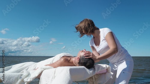 Female massage therapist does face massage to brunette woman on the summer beach, sea background. Relaxation from stress, relieve headache, health spa, lifestyle and body care, rehabilitation concept