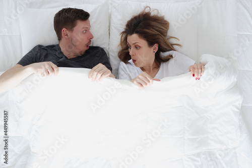 Bilde på lerret Surprised man and woman looking under covers while lying in bed