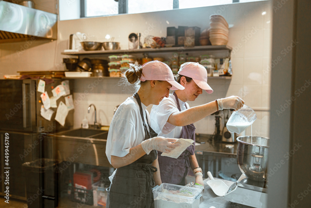 Employees pours milk into bowl making dough with colleague in craft bakery Employees pours milk into bowl making dough with colleague in craft bakery