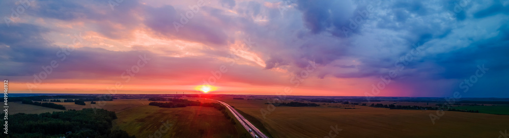 Panoramic aerial view of highway on red sunset. Landscape with road near countryside fields. Beautiful winding road leading through rural countryside with evening sunlight. Dramatic sky background.