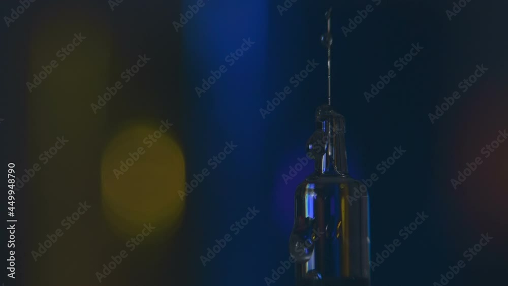 Drops of medicine dripping down the needle of a glass modern syringe for vaccine injection that glitters in colored light on a colorful background. Closeup. Macro. Slow mo, slo mo, slow motion, high s Drops of medicine dripping down the needle of a glass modern syringe for vaccine injection that glitters in colored light on a colorful background. Closeup. Macro. Slow mo, slo mo, slow motion, high s