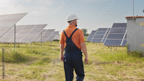 Maintenance assistance technical worker in uniform is checking an operation and efficiency performance of photovoltaic solar panels. Construction engineer walks between solar panels on field station.