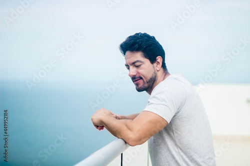 Shallow focus of a bearded Hispanic man leaning on a view deck railing with the sea in thebackground photo