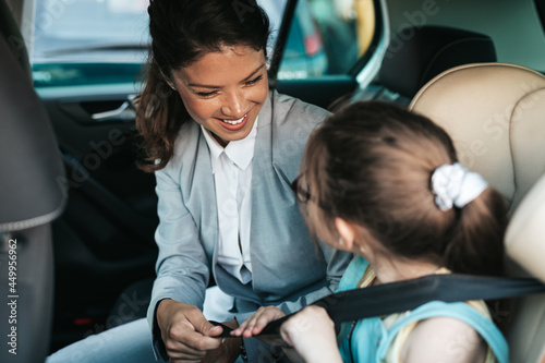 Young businesswoman helping her daughter to fasten seatbelts in the car while girl is sitting on a safety child car seat.