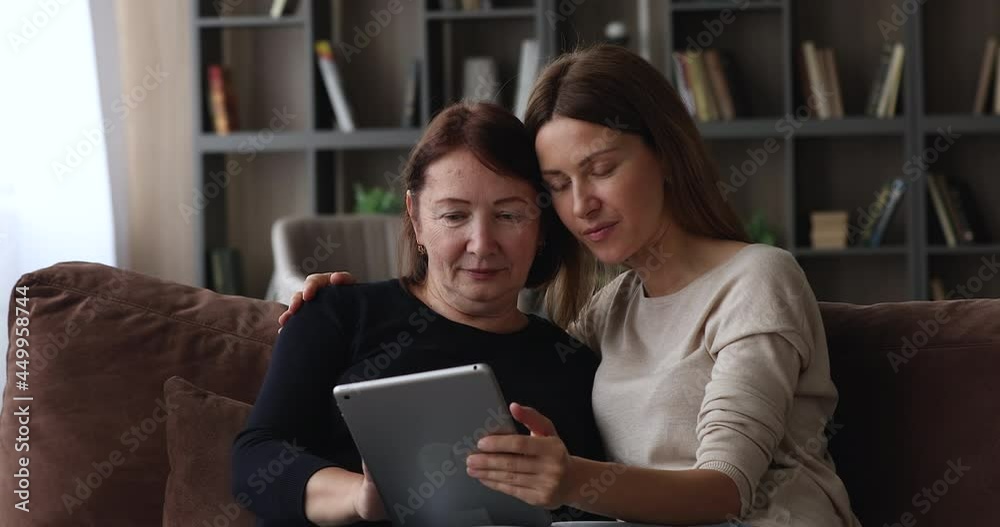 Caring smiling young woman teaching happy elderly senior retired mother using digital tablet software applications, shopping in internet store, playing or web surfing resting together on cozy sofa. Caring smiling young woman teaching happy elderly senior retired mother using digital tablet software applications, shopping in internet store, playing or web surfing resting together on cozy sofa.
