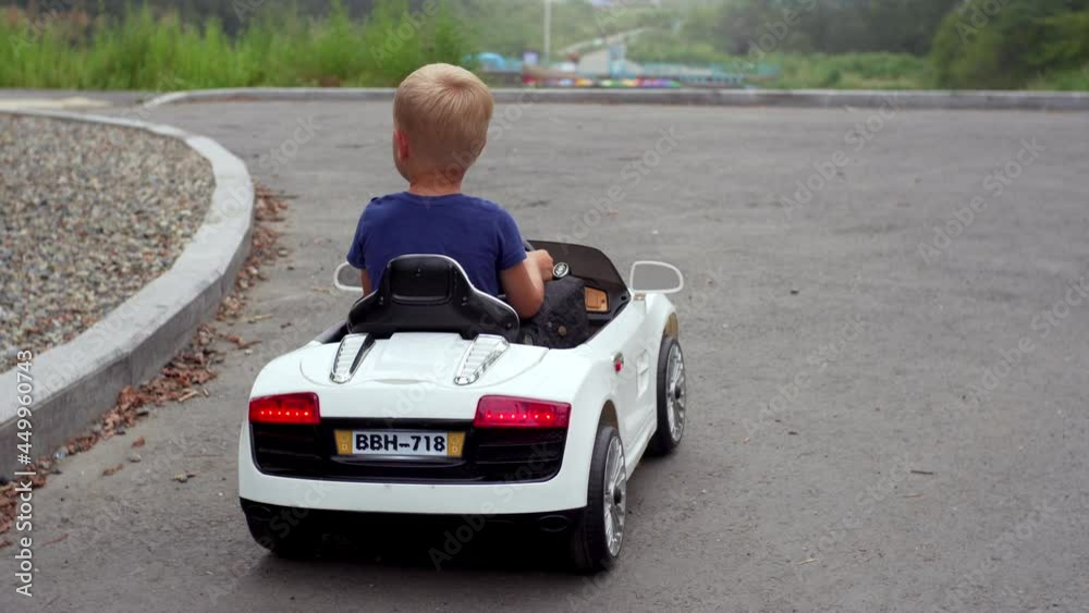Back of a 4 years old boy turning right in a white toy battery car in the summer park along the road. Summer activity with electric car in the park