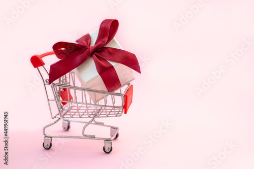 Shopping trolley with white gift box and red bow on pink background. Holiday shopping concept