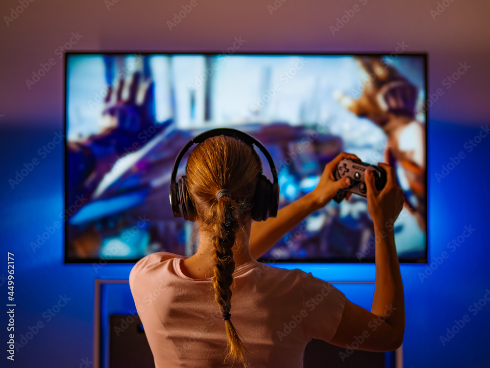 Girl in headphones plays a video game on the big TV screen. Gamer with a  joystick.