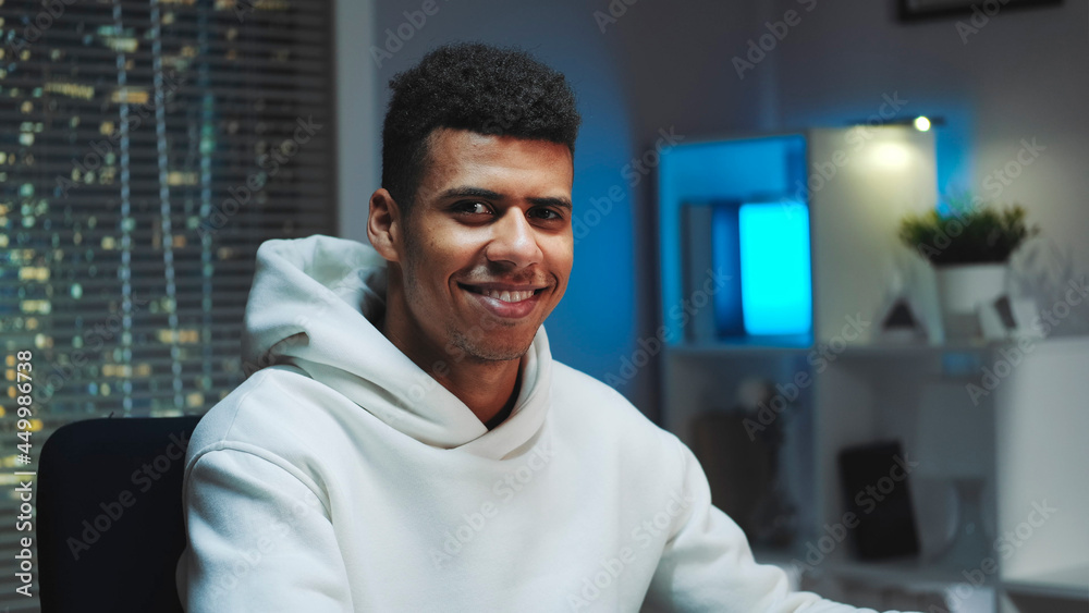 Close-up shot of Handsome black man smiling to the camera while working home on computer. Skyscrapers in the background. Close-up shot of Handsome black man smiling to the camera while working home on computer. Skyscrapers in the background.