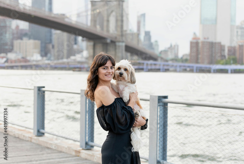 a woman holding her small mixed breed dog at the boardwalk by the hudson river with brooklyn bridge and new york city buildings in the background