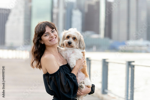 a woman holding her small mixed breed dog at the boardwalk by the hudson river with brooklyn bridge and new york city buildings in the background photo