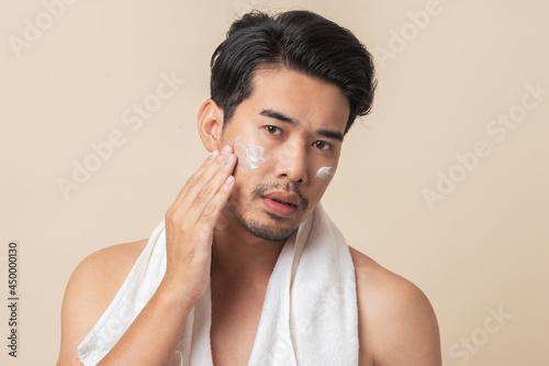 Young Asian man applying moisturizer skin care cream on face after take a shower on brown background. Asian man with mustaches. Men's skin care.