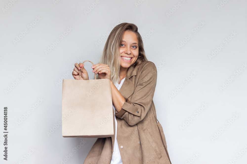 A young beautiful caucasian smiling blonde woman in a brown coat holds and demonstrates paper eco bag with copy empty space for the logo isolated on a light gray background.Shopping and fashion.Mockup A young beautiful caucasian smiling blonde woman in a brown coat holds and demonstrates paper eco bag with copy empty space for the logo isolated on a light gray background.Shopping and fashion.Mockup