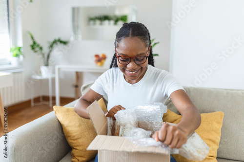 Smiling african American millennial girl sit on sofa at home open cardboard delivery package shopping online, excited black young woman unpack unwrap parcel buying good via internet or web