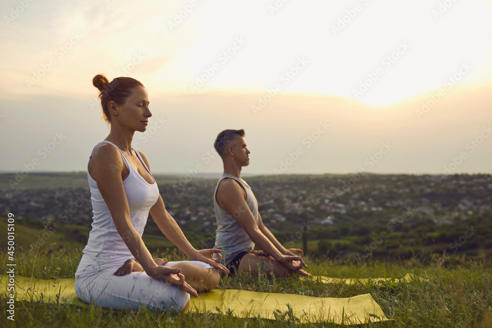 Man and woman meditating in Lotus position. Yogi couple sitting cross legged on green grass breathing deeply enjoying peace and quiet of nature at sunset. Yoga coach and student relaxing in Padmasana