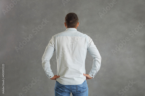 View from behind of athletic broad shouldered European man standing hands on hips against grey background. Studio backside shot of young guy with short hair in light blue denim shirt with mock up back