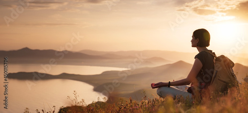 A young Asian woman with a backpack Sitting on a hill slope and enjoying the view of the sunset sea and mountains and meditates with a beautiful view . © YURII Seleznov