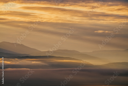 Scenic dawn mountain landscape with golden low clouds in valley among mountains silhouettes under cloudy sky. Vivid sunset or sunrise scenery with low clouds in mountain valley in illuminating color.
