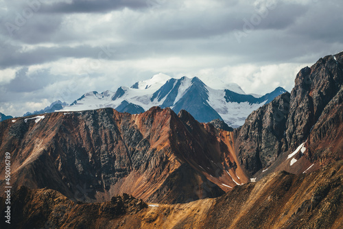 Atmospheric mountains landscape with great snowy top behind colorful brown red orange rocky wall under cloudy sky. Dramatic highland scenery with giant glacier and big vivid brown red orange mountain.