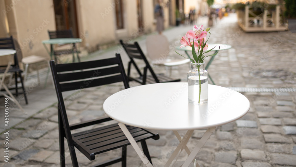 Beautiful delicate pink flower in a transparent glass vase on a white street table. Street view with cafe terrace in the morning in the city Beautiful delicate pink flower in a transparent glass vase on a white street table. Street view with cafe terrace in the morning in the city