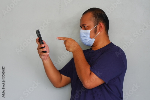 Young asian man wearing medical mask and pointing finger to smartphone