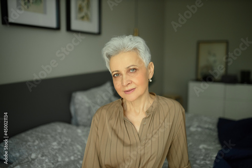 Lovely middle-aged woman with stylish grey haircut and healthy face with some wrinkles, wearing earrings, smiling confidently sitting on bed at home looking at camera. Aging gracefully concept