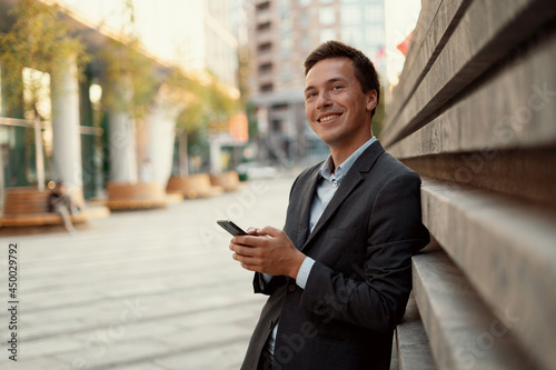 The manager is standing near the office writing a message to the client. He looks away and smiles. Business suit for a meeting.