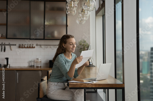 Happy young woman reading paper letter at home workplace with laptop, getting good news, bank or insurance notice. Gen Z student girl receiving approved application from college or university