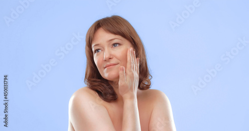 Pretty mature female applies skin care product by touching face with hand. Woman applying anti-age cream on her skin isolated on color background. Spa and wellness concept.