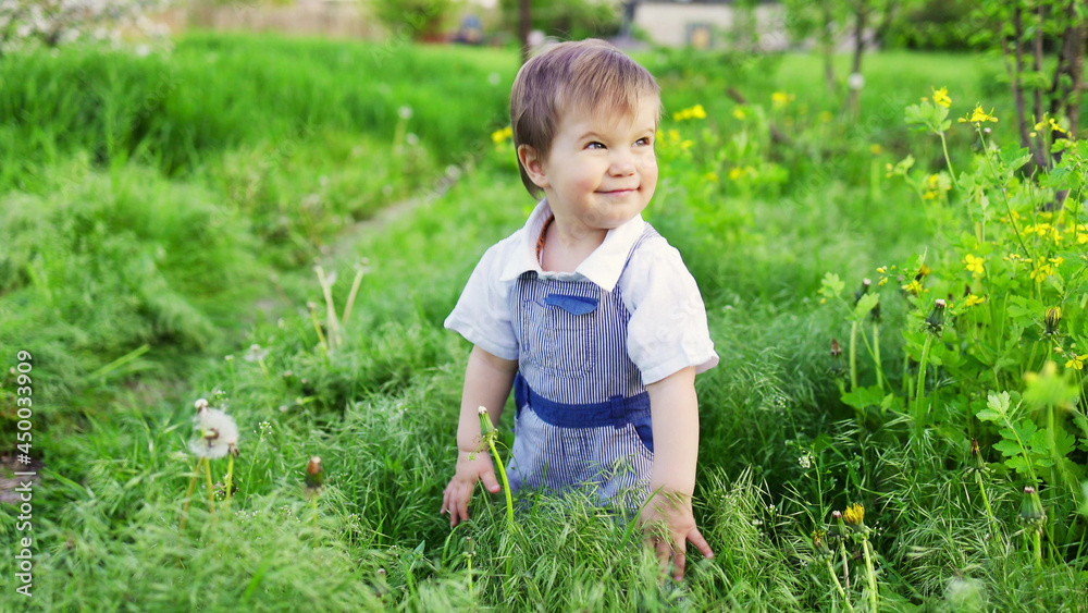 Cute boy in fashionable clothes with blue eyes plays and jumps in the tall grass in a large green blooming garden Cute boy in fashionable clothes with blue eyes plays and jumps in the tall grass in a large green blooming garden