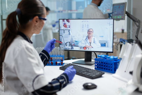 Biologist researcher woman holding blood test tube in hands discussing medical vaccine with chemist doctor during online videocall teleconference meeting in hospital lab. Scientist virtual conference