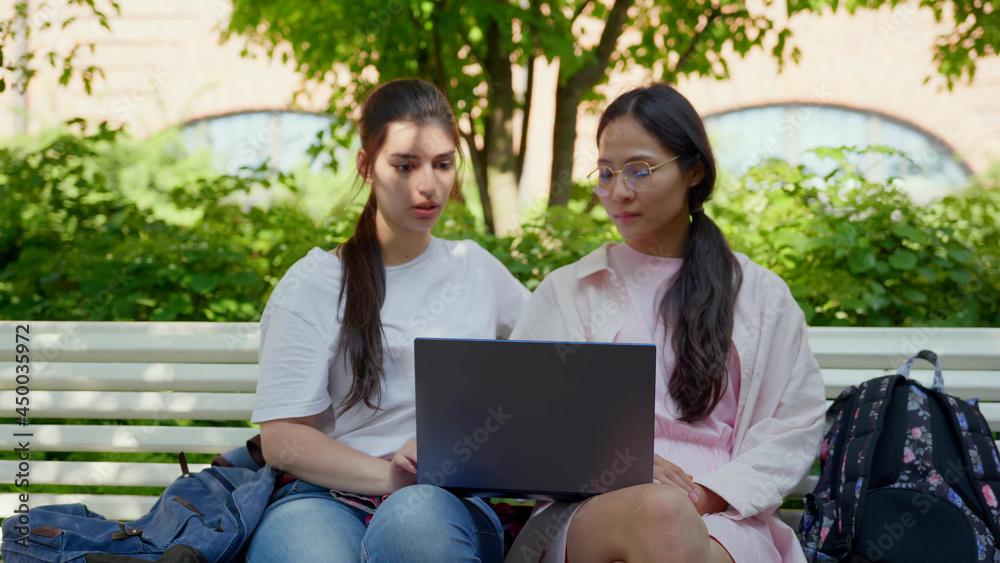 Young female students sitting on bench in park using laptop Young female students sitting on bench in park using laptop
