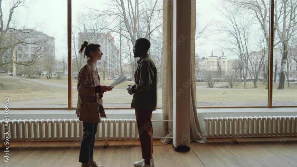 Zoom in shot of Afro-American man and Caucasian woman standing together in hall with floor-to-ceiling windows and discussing papers Zoom in shot of Afro-American man and Caucasian woman standing together in hall with floor-to-ceiling windows and discussing papers