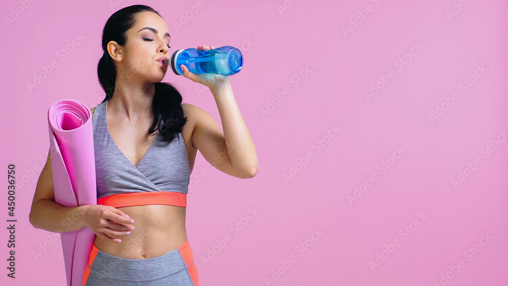young sportswoman drinking water from sports bottle and holding fitness mat isolated on pink young sportswoman drinking water from sports bottle and holding fitness mat isolated on pink