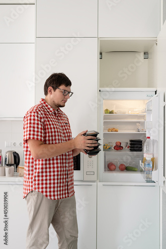 Young man holding disposable plastic boxed with food and putting them to the fridge