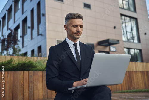 Confident mature businessman working on laptop while sitting outdoors near office building