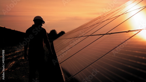 Assistance technical worker in uniform is checking an operation and efficiency performance of photovoltaic solar panels. Unidentified solar power engineer touches solar panels with his hand at sunset