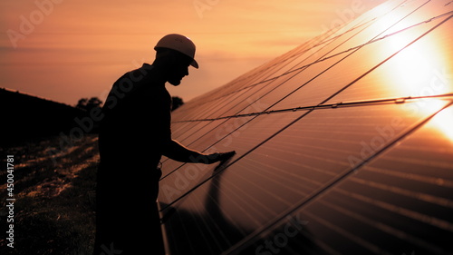 Assistance technical worker in uniform is checking an operation and efficiency performance of photovoltaic solar panels. Unidentified solar power engineer touches solar panels with his hand at sunset