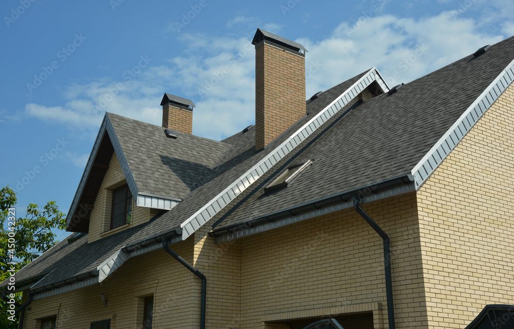 A close-up of a double complex roof of a brick house construction covered with gray asphalt shingles, fascia and soffit, with an attic skylight window, chimneys, roof gutters and downspouts.