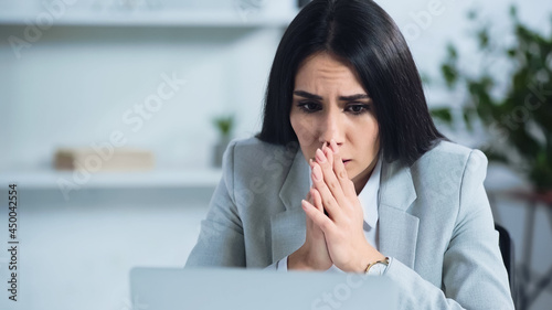 worried businesswoman with praying hands near blurred laptop in office