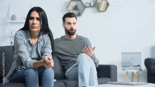 Fototapeta Naklejka Na Ścianę i Meble -  worried woman sitting with clenched hands near blurred and displeased boyfriend