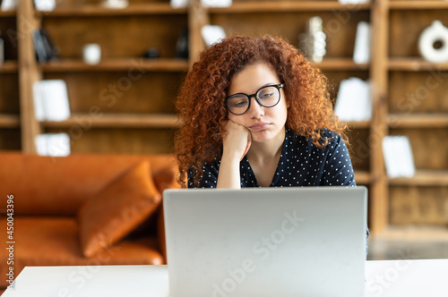 Young redhead curly woman using laptop sitting in the office and feels bored, fatigue, lack of motivation and energy, female employee tired from not interesting and monotonous work photo