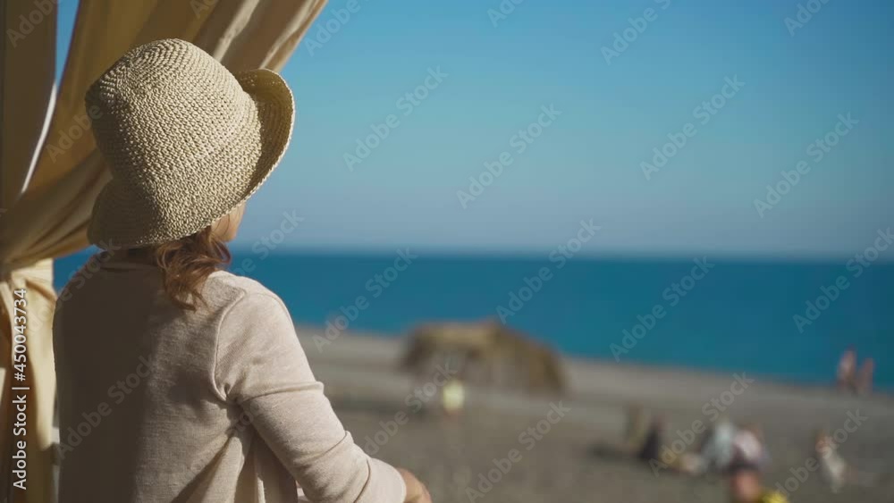 Beautiful young girl with a straw hat looks from the balcony at the blue sea. Rear view from the back. Natural style in clothing and interior. Concept of rest, travel, vacation, magazine, relaxation Beautiful young girl with a straw hat looks from the balcony at the blue sea. Rear view from the back. Natural style in clothing and interior. Concept of rest, travel, vacation, magazine, relaxation