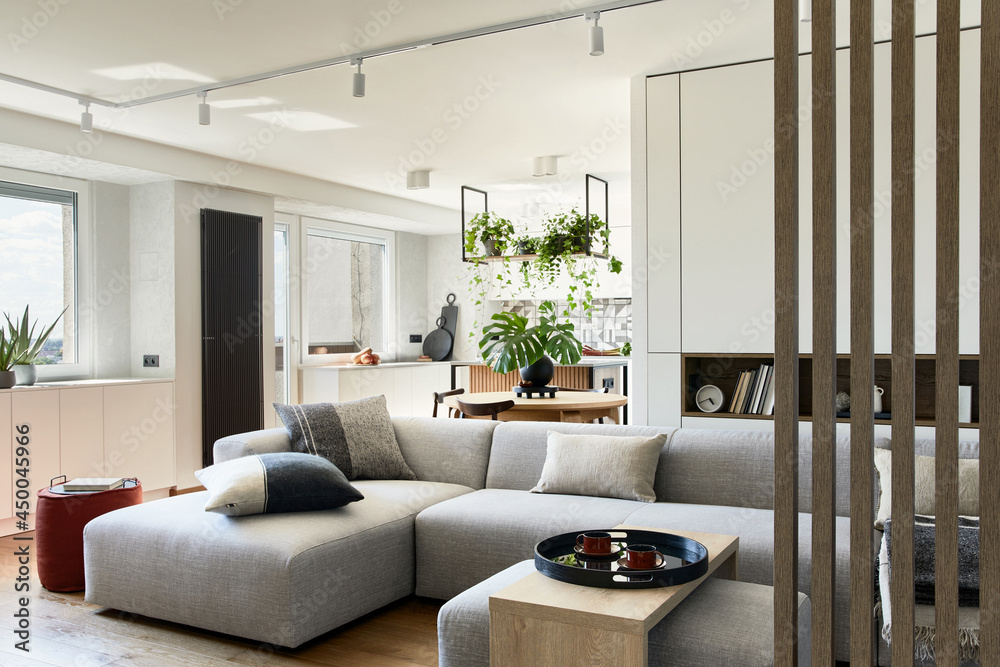 Stylish living room interior design with grey sofa, pouf and personal accessories. Dining space and kitchen on the background. Creative walls with woode pannels. Minimalistic style concept. Stylish living room interior design with grey sofa, pouf and personal accessories. Dining space and kitchen on the background. Creative walls with woode pannels. Minimalistic style concept.