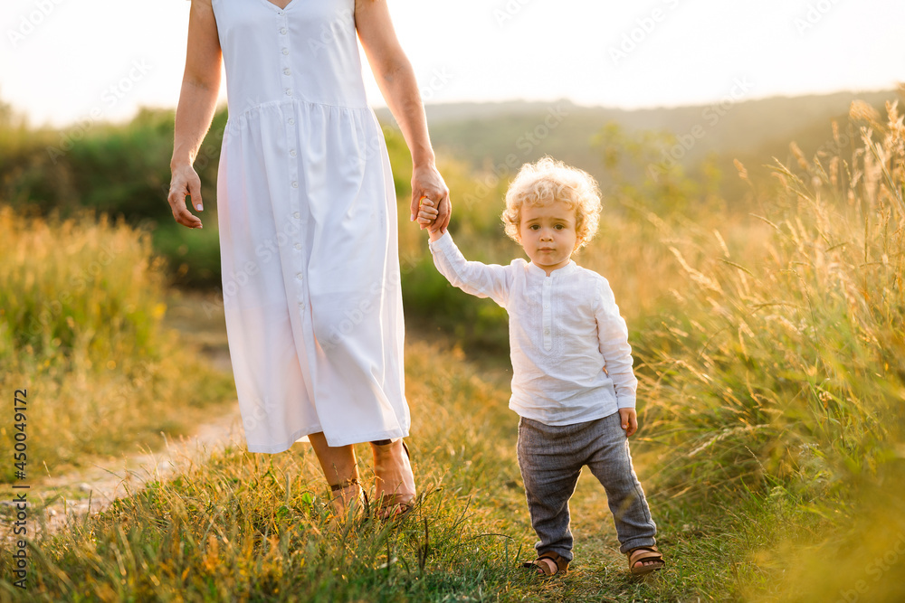Fototapeta premium Beautiful woman in light white dress leads her son by the hand along the path of a picturesque field