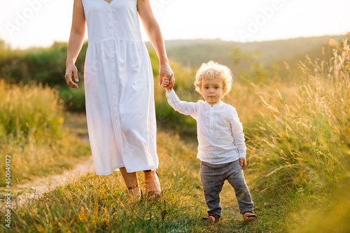 Beautiful woman in light white dress leads her son by the hand along the path of a picturesque field