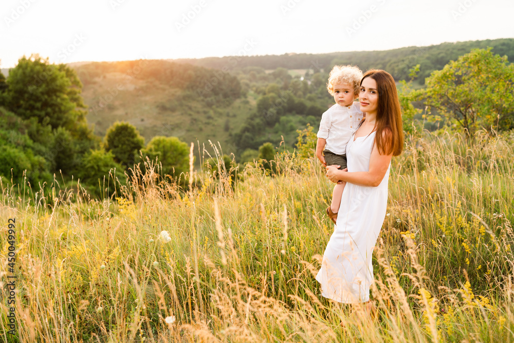 Portrait of a beautiful woman in a white dress with a cute curly boy in her arms in nature