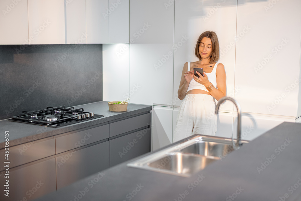Young woman dressed in white standing relaxed with phone on the modern kitchen at home. Smart home and modern living concept Young woman dressed in white standing relaxed with phone on the modern kitchen at home. Smart home and modern living concept