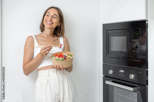 Happy woman eating asian takeaway food heated in microwave machine at home. Concept of modern and heallthy life with modern kitchen appliances photo