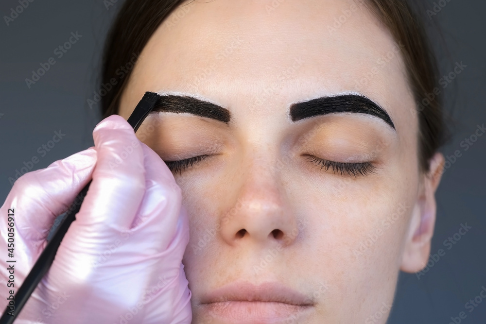 Cosmetologist is painting woman's eyebrows in cosmetology clinic, closeup face, front view. Applying brown paint on eyebrows into white contour using brush. Beauty procedure for woman in beauty salon.